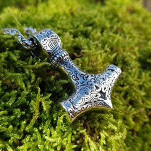 Op de afbeelding: Een zilveren Mjolnir-hanger met ingewikkelde bloemmotieven, hangend aan een zilveren ketting. De hanger ligt op een bed van groen mos.
