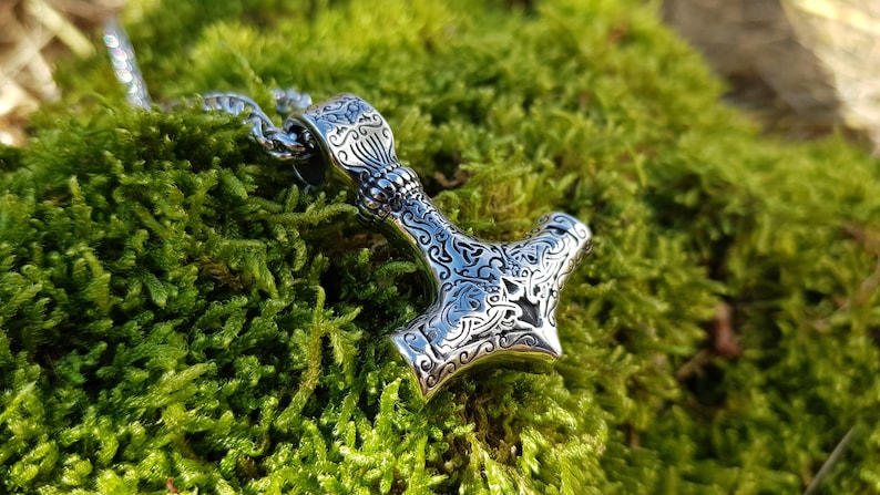 Op de afbeelding: Een zilveren Mjolnir-hanger met ingewikkelde bloemmotieven, hangend aan een zilveren ketting. De hanger ligt op een bed van groen mos.