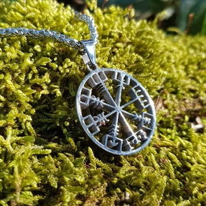 Puede incluir: Un collar de plata con un colgante circular que presenta un dise&ntilde;o de rosa de los vientos con runas. El colgante est&aacute; sobre un lecho de musgo verde.