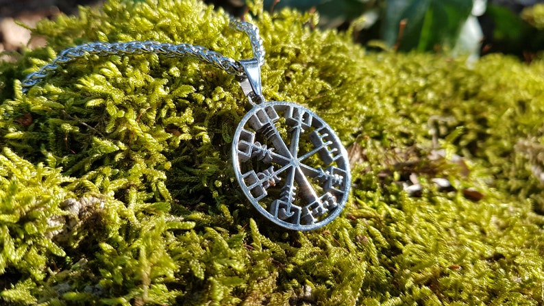 Puede incluir: Un collar de plata con un colgante circular que presenta un dise&ntilde;o de rosa de los vientos con runas. El colgante est&aacute; sobre un lecho de musgo verde.