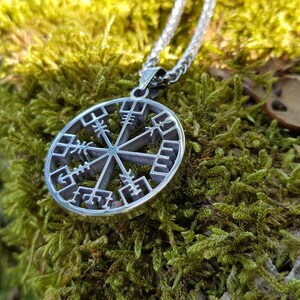 Puede incluir: Un collar de plata con un colgante circular que presenta un s&iacute;mbolo Vegvisir, una br&uacute;jula direccional n&oacute;rdica, sobre un fondo de musgo verde.