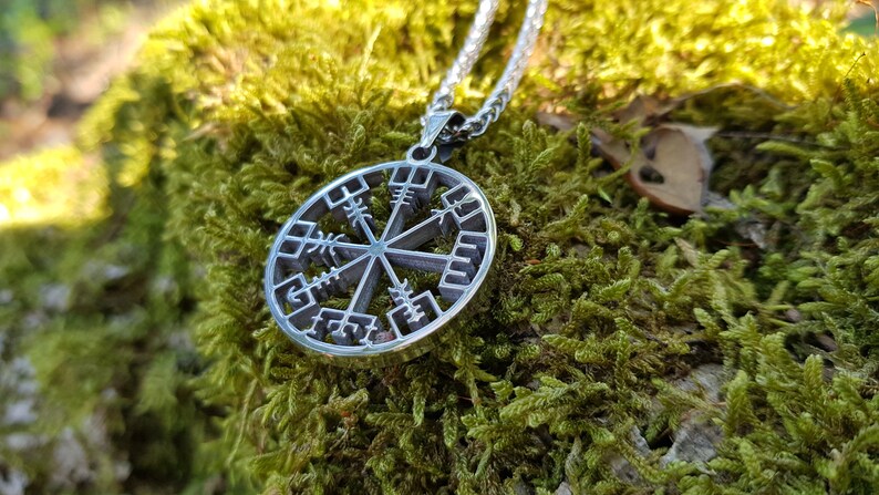Puede incluir: Un collar de plata con un colgante circular que presenta un s&iacute;mbolo Vegvisir, una br&uacute;jula direccional n&oacute;rdica, sobre un fondo de musgo verde.