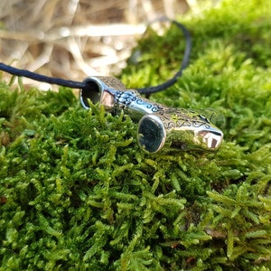 Op de afbeelding: Zilveren ketting met een gedetailleerd ontwerp van een hamer, op een zwart koord, liggend op een bed van groen mos.