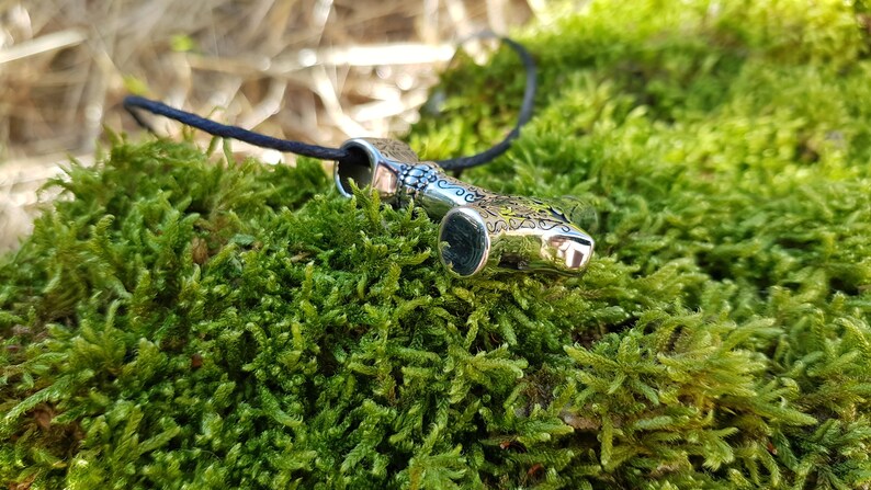 Op de afbeelding: Zilveren ketting met een gedetailleerd ontwerp van een hamer, op een zwart koord, liggend op een bed van groen mos.