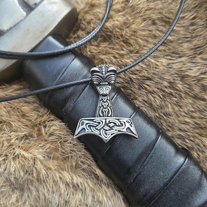 Könnte beinhalten: Eine silberfarbene Halskette mit Anhänger, die Thors Hammer, Mjolnir, zeigt. Der Anhänger weist komplizierte keltische Knotenmuster auf und hängt an einer schwarzen Kordel. Die Halskette liegt auf einer strukturierten braunen Oberfläche.
