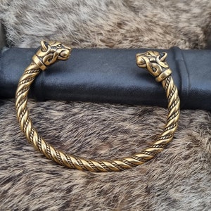 May include: A gold-toned braided bracelet with wolf head terminals. The bracelet is lying on a brown fur surface.