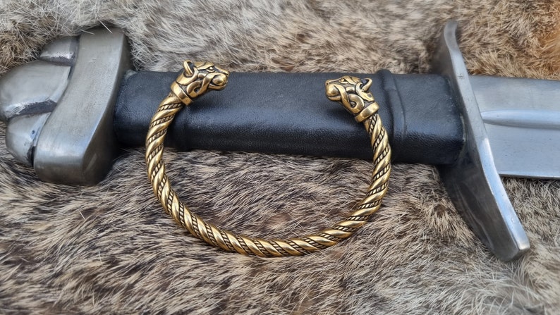 May include: A gold-toned braided bracelet with wolf head terminals. The bracelet is lying on a brown fur surface.