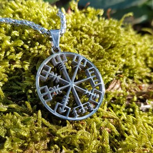 Puede incluir: Un collar de plata con un colgante circular que presenta un s&iacute;mbolo Vegvisir, una br&uacute;jula n&oacute;rdica, con detalles intrincados. El collar est&aacute; descansando sobre un lecho de musgo verde.