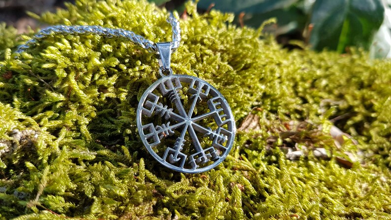 Puede incluir: Un collar de plata con un colgante circular que presenta un s&iacute;mbolo Vegvisir, una br&uacute;jula n&oacute;rdica, con detalles intrincados. El collar est&aacute; descansando sobre un lecho de musgo verde.
