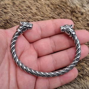 May include: A silver braided bracelet with wolf head terminals. The bracelet is on a furry surface.