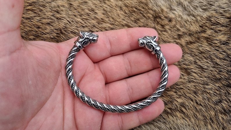 May include: A silver braided bracelet with wolf head terminals. The bracelet is on a furry surface.