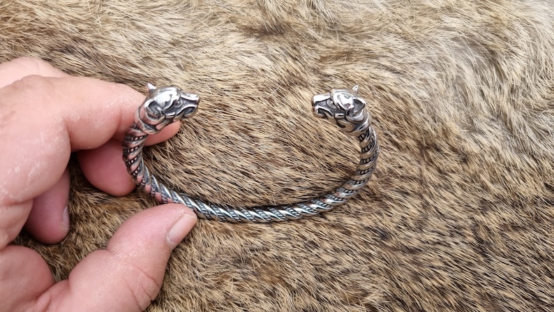 May include: A silver bracelet with two wolf head ends. The bracelet is twisted and has a braided design.