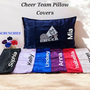 May include: Set of 7 satin pillow covers with the words "Cheer Team Pillow Covers" at the top. The pillow covers are in various colours, including black, purple, blue, white, and pink. Each pillow cover has a name embroidered on it, including Sophia, Kelly, Lindsey, Anna, Isabella, Piper, and Mia. The pillow cover with Mia's name has a cheerleading megaphone and a paw print embroidered on it.