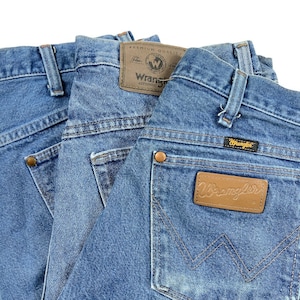 May include: Three pairs of blue denim jeans with a brown leather Wrangler label on the back pocket. The label says "Wrangler Authentic Premium Quality".