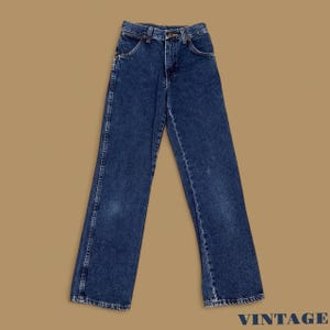 Puede incluir: Vaqueros de mezclilla azul oscuro con un diseño clásico de cinco bolsillos. Los vaqueros tienen un corte de pierna recta y un cierre de botón y cremallera. La palabra "VINTAGE" está impresa en azul marino en la esquina inferior derecha.