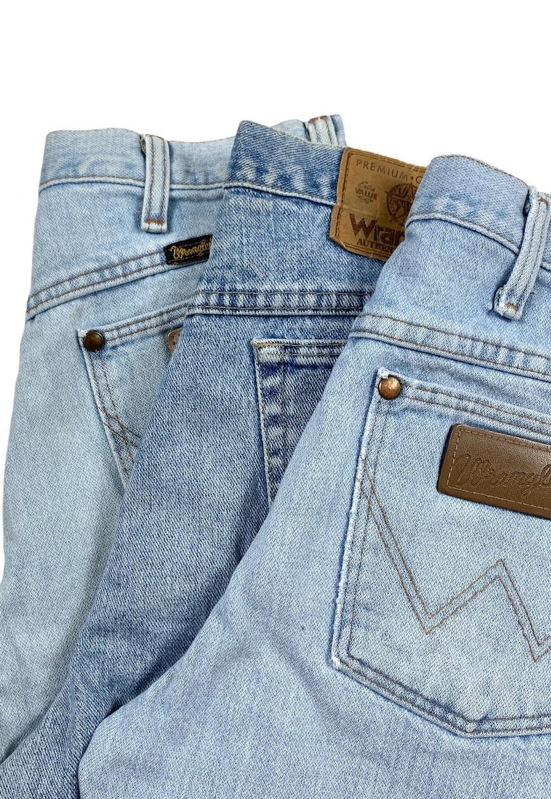 Peut inclure: Gros plan sur trois paires de jeans en denim bleu clair. Les jeans ont une coupe classique &agrave; cinq poches, avec des coutures visibles et des rivets cuivr&eacute;s. L'&eacute;tiquette de la marque Wrangler est visible sur la poche arri&egrave;re.