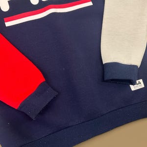 May include: A navy blue sweatshirt with red and gray sleeves. The sweatshirt features the brand name in white with a red stripe. The cuffs and hem are navy blue. The brand label is visible on the gray sleeve.