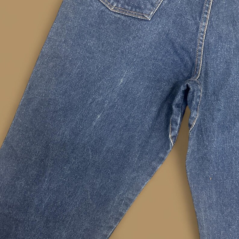 K&ouml;nnte beinhalten: Nahaufnahme einer blauen Jeans. Die Jeans weist Gebrauchsspuren auf, mit sichtbaren Ausbleichungen und hellen Flecken. Die N&auml;hte sind entlang der N&auml;hte und Taschen sichtbar. Der Gesamteindruck ist l&auml;ssig und vintage.