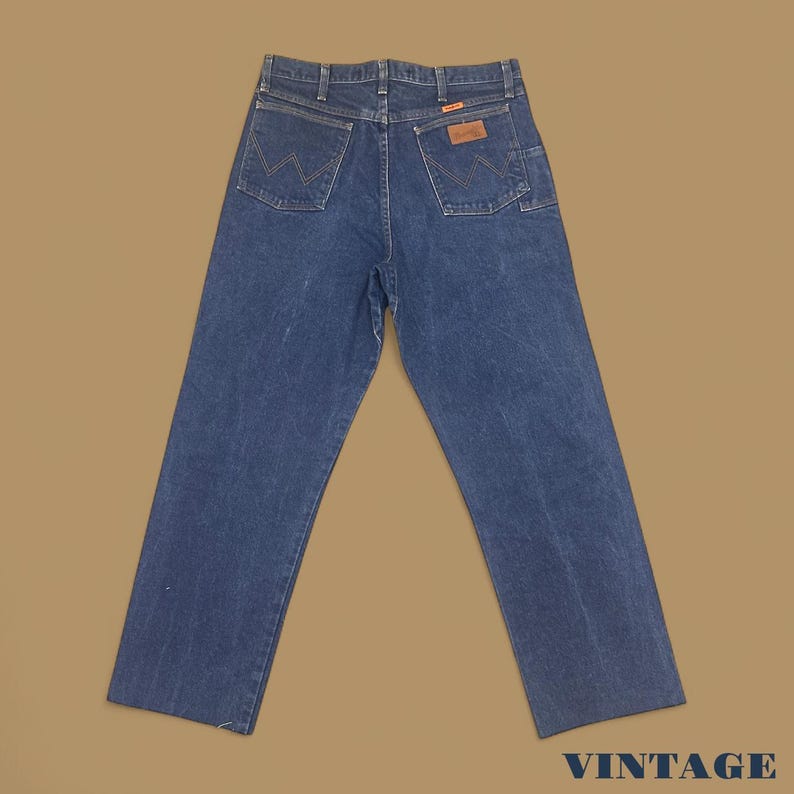 K&ouml;nnte beinhalten: Dunkelblaue Jeans mit dem Wrangler-Logo auf den Ges&auml;&szlig;taschen. Die Jeans haben ein klassisches F&uuml;nf-Taschen-Design und einen braunen Lederaufn&auml;her. Das Wort "VINTAGE" ist unten aufgedruckt.