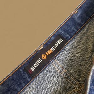 K&ouml;nnte beinhalten: Nahaufnahme einer blauen Jeans mit einem Relaxed Fit-Etikett und dem Text Flame Resistant. Das Etikett ist schwarz mit wei&szlig;em und rotem Text. Die Jeans hat ein klassisches F&uuml;nf-Taschen-Design und ist aus strapazierf&auml;higem Denim gefertigt.