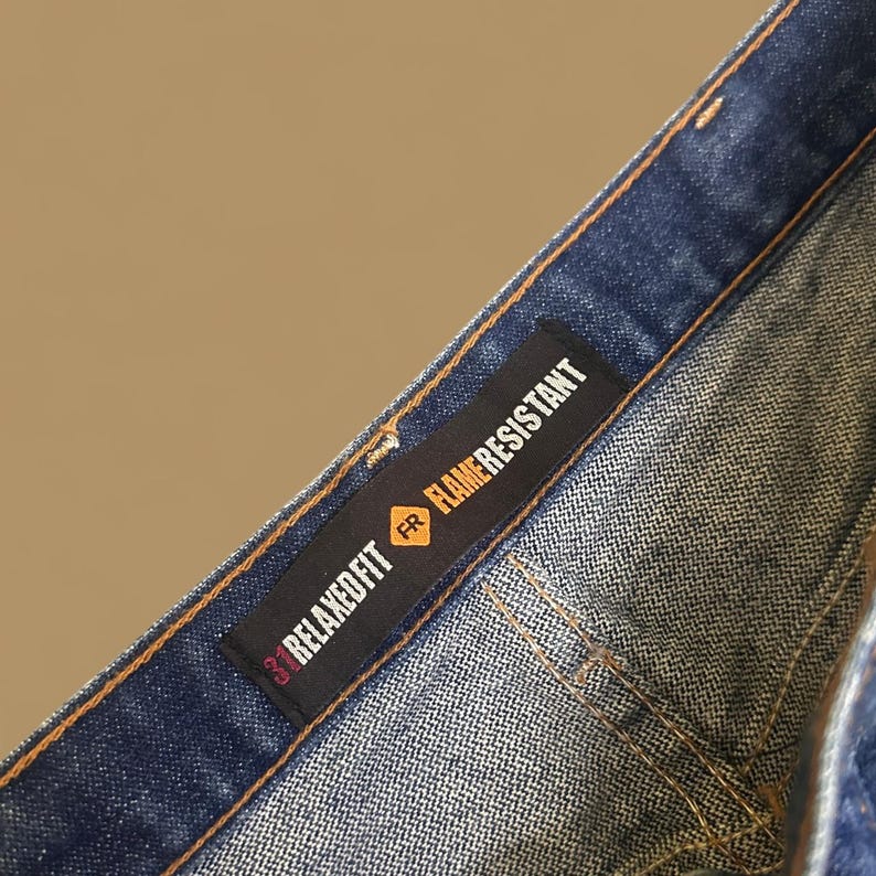 K&ouml;nnte beinhalten: Nahaufnahme einer blauen Jeans mit einem Relaxed Fit-Etikett und dem Text Flame Resistant. Das Etikett ist schwarz mit wei&szlig;em und rotem Text. Die Jeans hat ein klassisches F&uuml;nf-Taschen-Design und ist aus strapazierf&auml;higem Denim gefertigt.