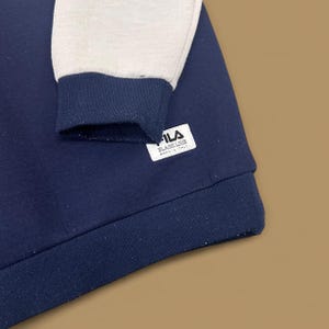 May include: A navy blue sweatshirt with light gray sleeves and a navy blue ribbed cuff. The sleeve cuff has a white tag with the text "FILA BLACK LINE BORN IN ITALY". The sweatshirt is made of a soft, comfortable material.