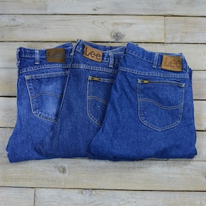 Puede incluir: Tres pares de pantalones vaqueros de mezclilla azul con etiquetas Lee de cuero marrón. Los pantalones vaqueros están doblados y apilados uno encima del otro.