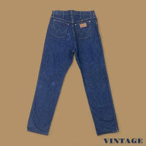 May include: Vintage blue denim jeans with a classic five-pocket design. The jeans feature a Wrangler brand patch on the back pocket and a faded appearance. The word "VINTAGE" is printed in the bottom right corner.
