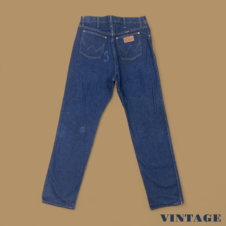 May include: Vintage blue denim jeans with a classic five-pocket design. The jeans feature a Wrangler brand patch on the back pocket and a faded appearance. The word "VINTAGE" is printed in the bottom right corner.