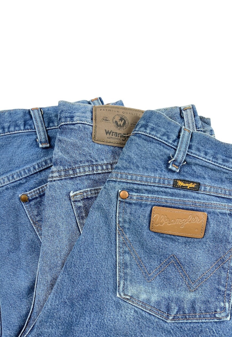 May include: Three pairs of blue denim jeans with a brown leather Wrangler label on the back pocket. The label says "Wrangler Authentic Premium Quality".