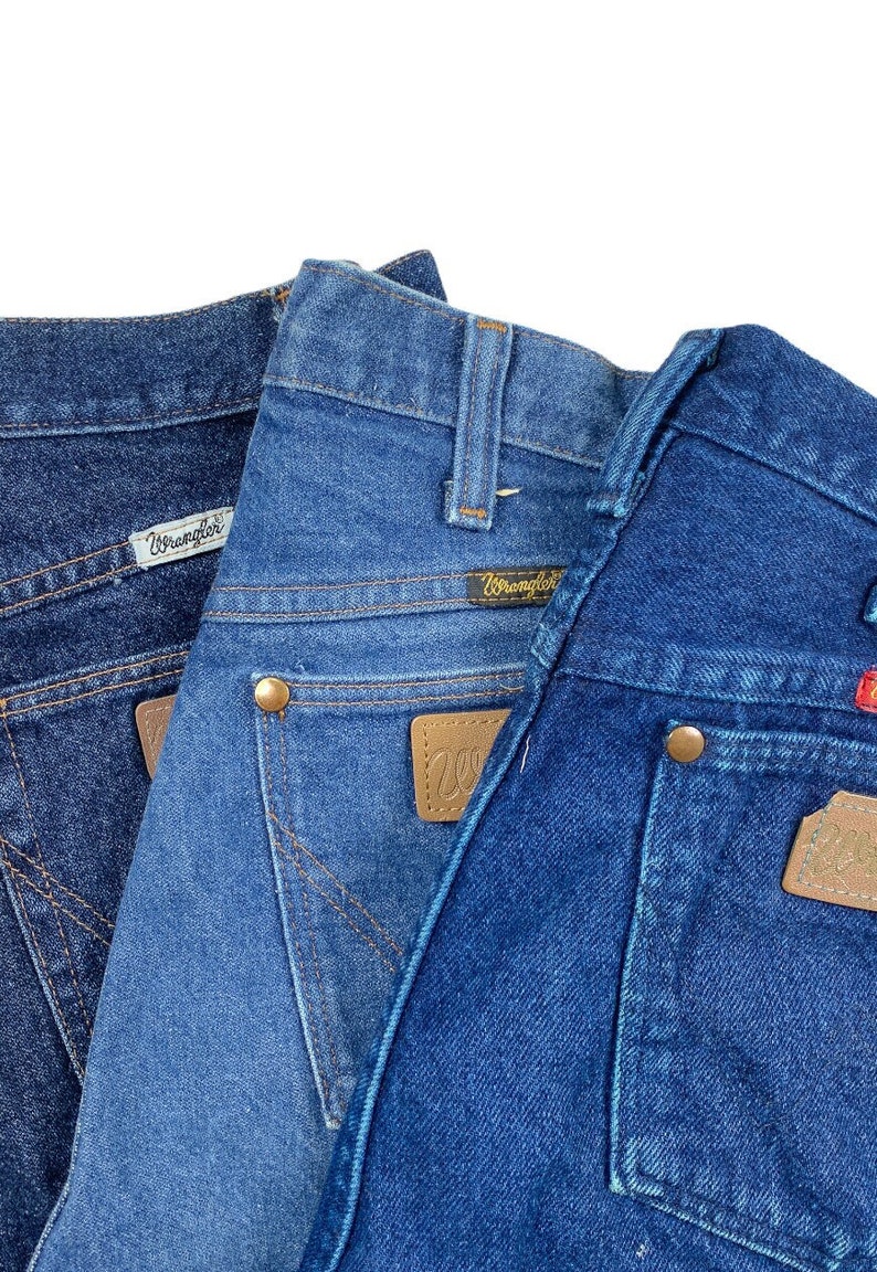K&ouml;nnte beinhalten: Zwei Paar blaue Jeans aus Denim mit einem braunen Leder-Wrangler-Patch auf der Ges&auml;&szlig;tasche. Die Jeans sind dunkelblau und hellblau.