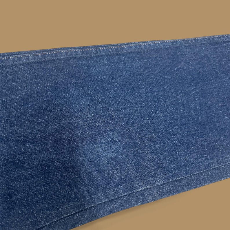 May include: Close-up of a section of blue denim jeans. The fabric shows a slight discoloration in the center. The stitching along the top edge is visible. The jeans are a medium wash.