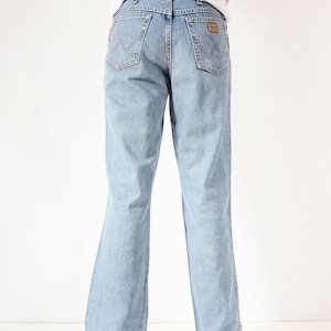 May include: A pair of light blue denim jeans with a straight leg fit. The jeans have a brown leather patch on the back with the Wrangler logo.