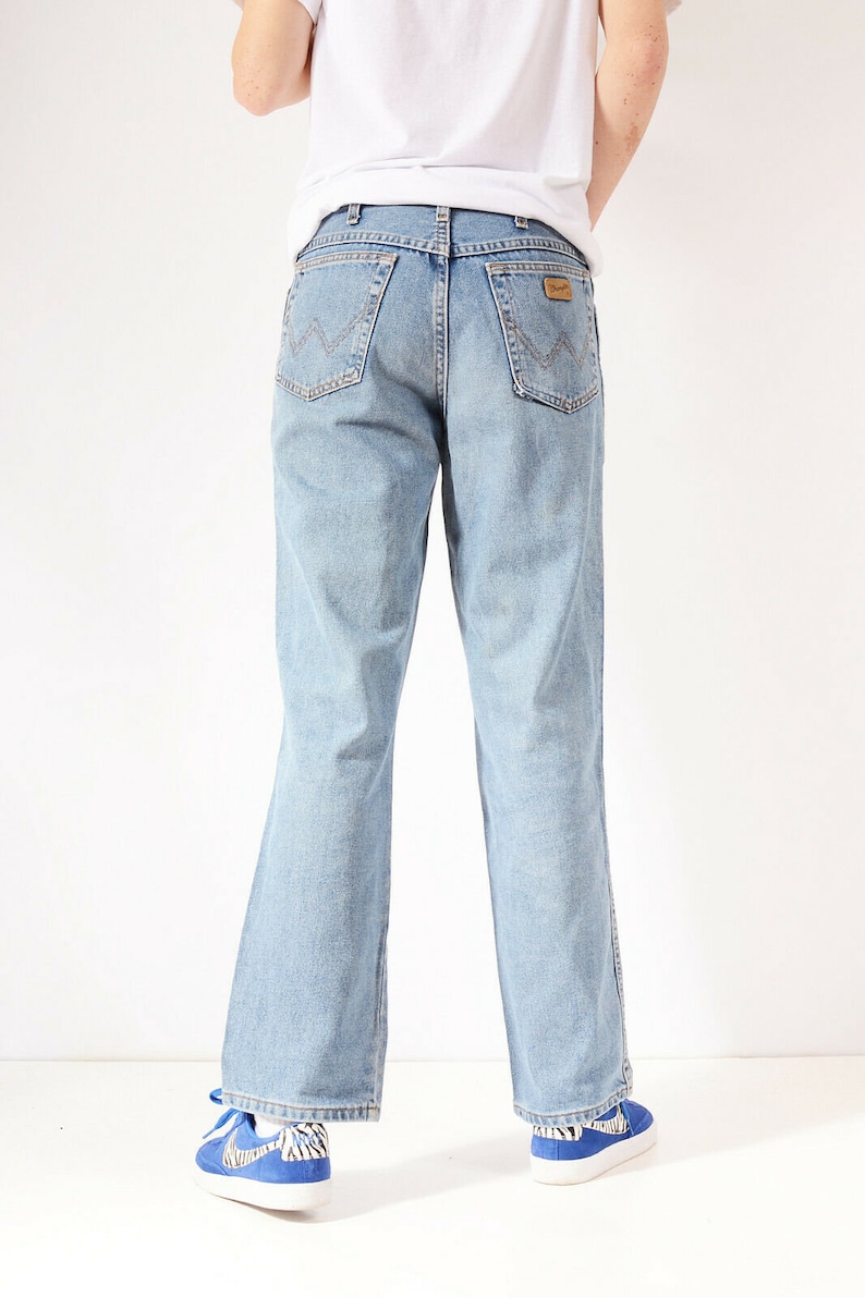 May include: A pair of light blue denim jeans with a straight leg fit. The jeans have a brown leather patch on the back with the Wrangler logo.