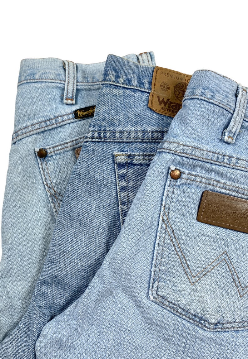 Puede incluir: Tres pares de pantalones vaqueros de mezclilla azul con diferentes bolsillos traseros y etiquetas. Los vaqueros tienen un lavado claro y est&aacute;n doblados uno encima del otro.