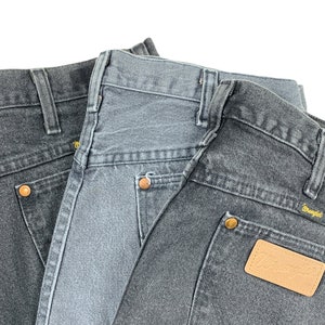 May include: Two pairs of dark gray denim jeans with a brown leather patch on the back pocket of the bottom pair. The patch has the word "Wrangler" printed on it.