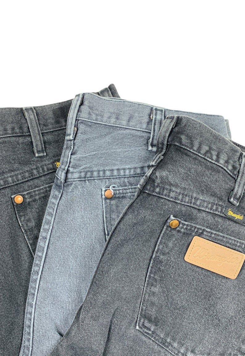 May include: Two pairs of dark gray denim jeans with a brown leather patch on the back pocket of the bottom pair. The patch has the word "Wrangler" printed on it.