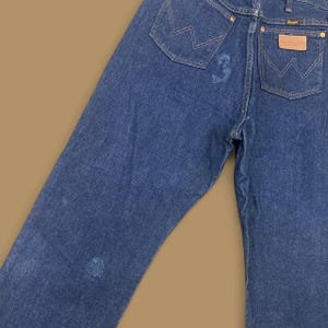 May include: Close-up of blue denim jeans with the Wrangler logo on the back pocket. The jeans have a classic five-pocket design and a faded appearance. The stitching is visible, and the jeans appear to be vintage.