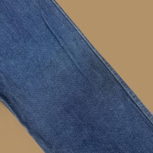 May include: Close-up of blue denim fabric. The denim has a classic, slightly faded appearance with visible texture and subtle variations in color. The image shows a portion of a garment, with a seam running diagonally across the frame.
