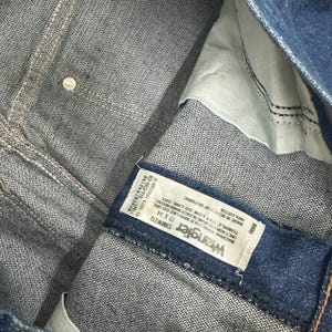 May include: Close-up of blue denim jeans with a white label that reads "Wrangler 13MWZG 17 X 34". The jeans are made of 100% cotton and are manufactured in Mexico.