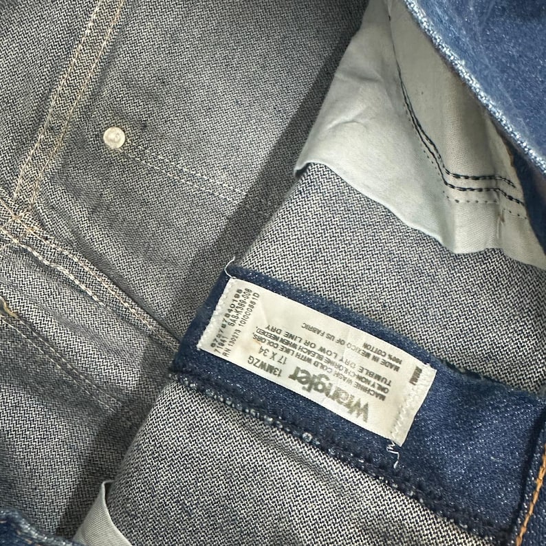 May include: Close-up of blue denim jeans with a white label that reads "Wrangler 13MWZG 17 X 34". The jeans are made of 100% cotton and are manufactured in Mexico.