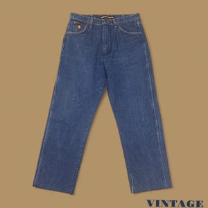 K&ouml;nnte beinhalten: Blaue Jeans aus Denim mit geradem Bein. Die Jeans haben ein klassisches F&uuml;nf-Taschen-Design, einen Knopf- und Rei&szlig;verschluss und ein kleines orangefarbenes Logo auf der Vordertasche. Das Wort "VINTAGE" ist unten rechts aufgedruckt.