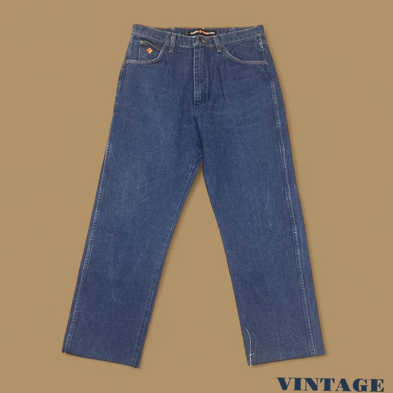 K&ouml;nnte beinhalten: Blaue Jeans aus Denim mit geradem Bein. Die Jeans haben ein klassisches F&uuml;nf-Taschen-Design, einen Knopf- und Rei&szlig;verschluss und ein kleines orangefarbenes Logo auf der Vordertasche. Das Wort "VINTAGE" ist unten rechts aufgedruckt.