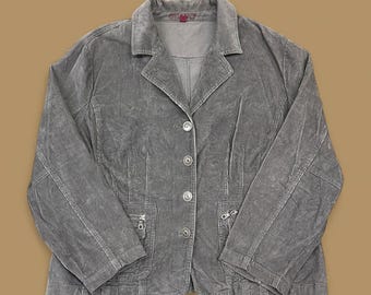 Op de afbeelding: Een grijze corduroy blazer met een reverskraag, knoopsluiting en twee voorzakken met ritsen. De blazer heeft lange mouwen en het woord "VINTAGE" staat rechtsonder gedrukt.