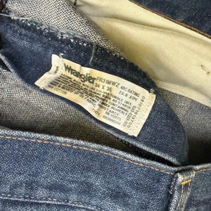 K&ouml;nnte beinhalten: Nahaufnahme einer blauen Wrangler-Jeans. Das Etikett lautet "FR31MWZ 34 X 36" mit Wasch- und Stoffinformationen. Die Jeans besteht aus 100 % Baumwolle.