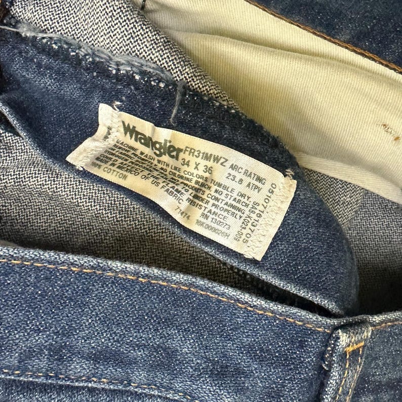 K&ouml;nnte beinhalten: Nahaufnahme einer blauen Wrangler-Jeans. Das Etikett lautet "FR31MWZ 34 X 36" mit Wasch- und Stoffinformationen. Die Jeans besteht aus 100 % Baumwolle.