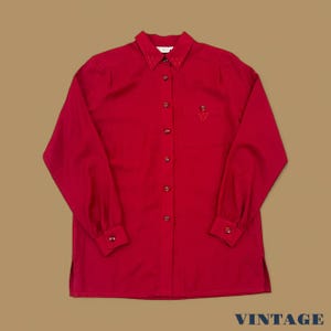 Peut inclure: Une chemise rouge à manches longues avec un col et une poche poitrine. La chemise a des poignets boutonnés et une petite broderie sur le col et la poche. Le mot "VINTAGE" est imprimé en bas.