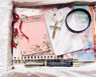 May include: A gift box containing a variety of items. Includes a pink notepad with floral accents and the text "Forgive and you will be forgiven", a cross keychain, a black bracelet, and a pen with the text "For with God nothing shall be impossible".