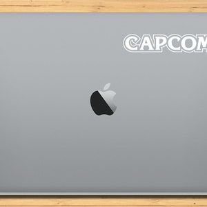 May include: A silver laptop computer with a white Apple logo and a white sticker that says "CAPCOM" on the top right corner.