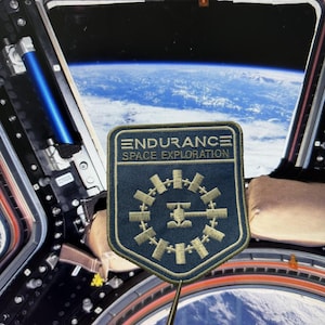 Op de afbeelding: Een donkerblauwe geborduurde patch met een gouden rand en gouden stiksels. De tekst ENDURANCE SPACE EXPLORATION staat boven een afbeelding van een cirkelvormig ruimtestation. De patch is geplaatst tegen een uitzicht op de aarde.